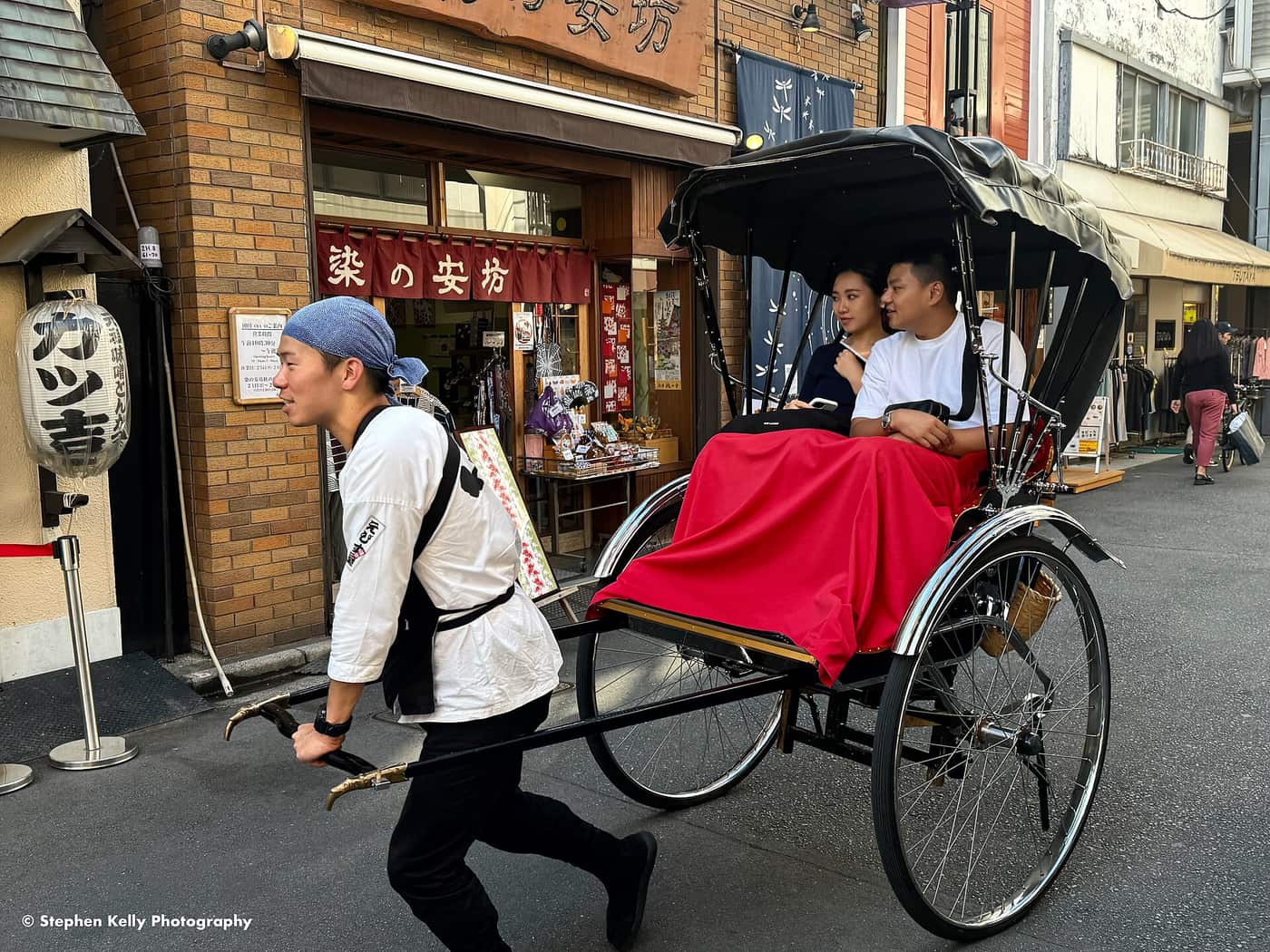 Rickshaw puller hauling two passengers past shopfronts on an Asakusa street