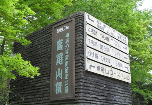 Summit sign of Mt Takao with trail direction boards among fresh green trees