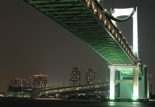 Underside of Rainbow Bridge glowing green at night over the waters of Odaiba